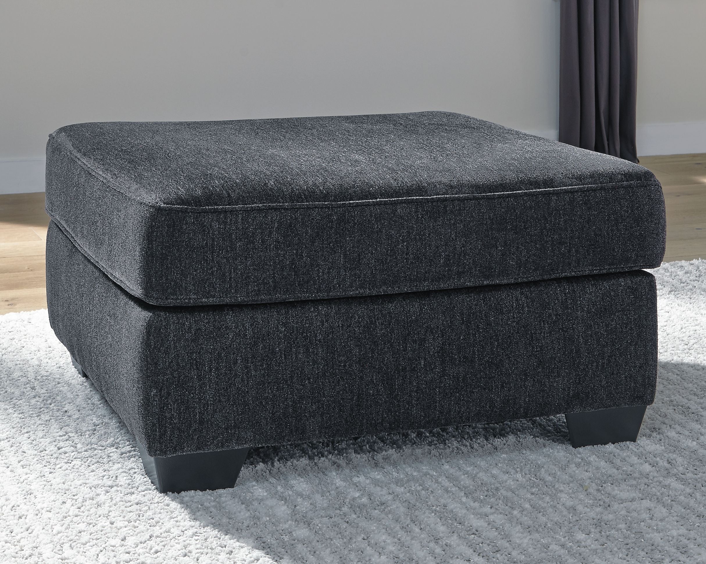 Dark gray upholstered accent ottoman with padded seat, rounded edges, and short legs