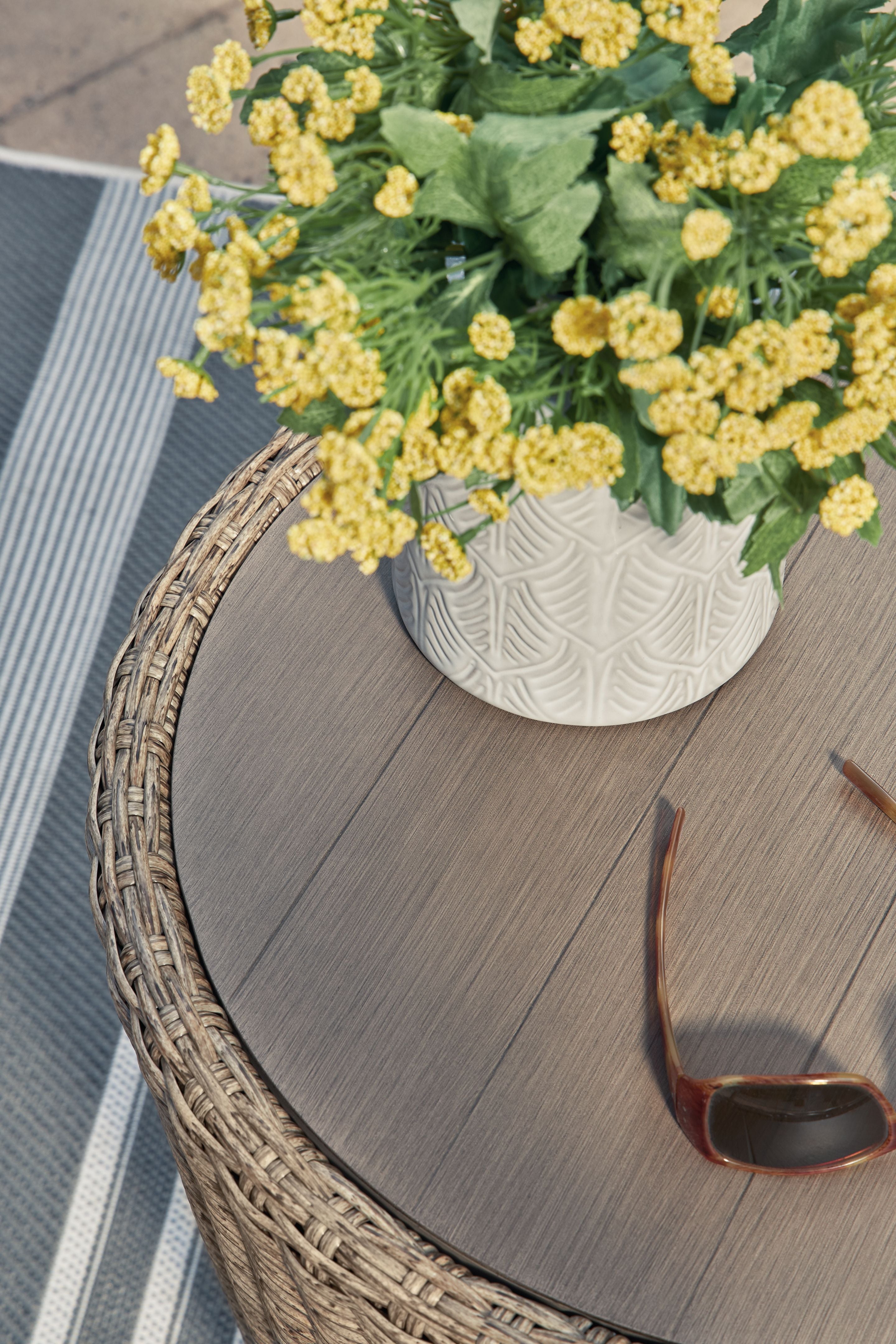 Beige round end table with woven rim and wood-tone tabletop with a white vase.