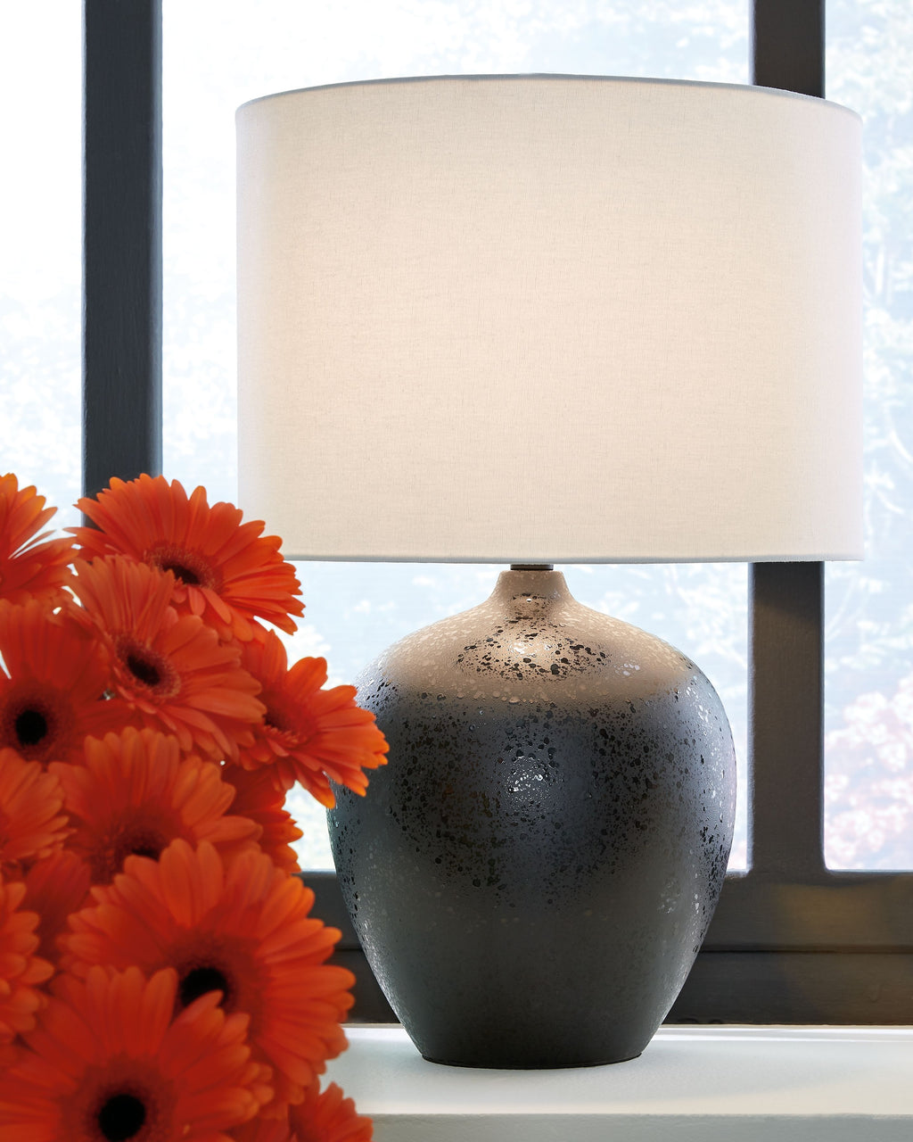 Ceramic table lamp with black speckled base and white drum shade near orange flowers