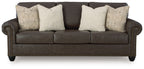 Dark brown sofa with four beige patterned pillows, rolled arms, and studded base