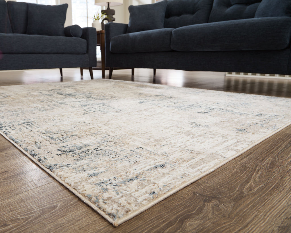 Cream abstract area rug with blue-gray distressed pattern placed between dark sofas.