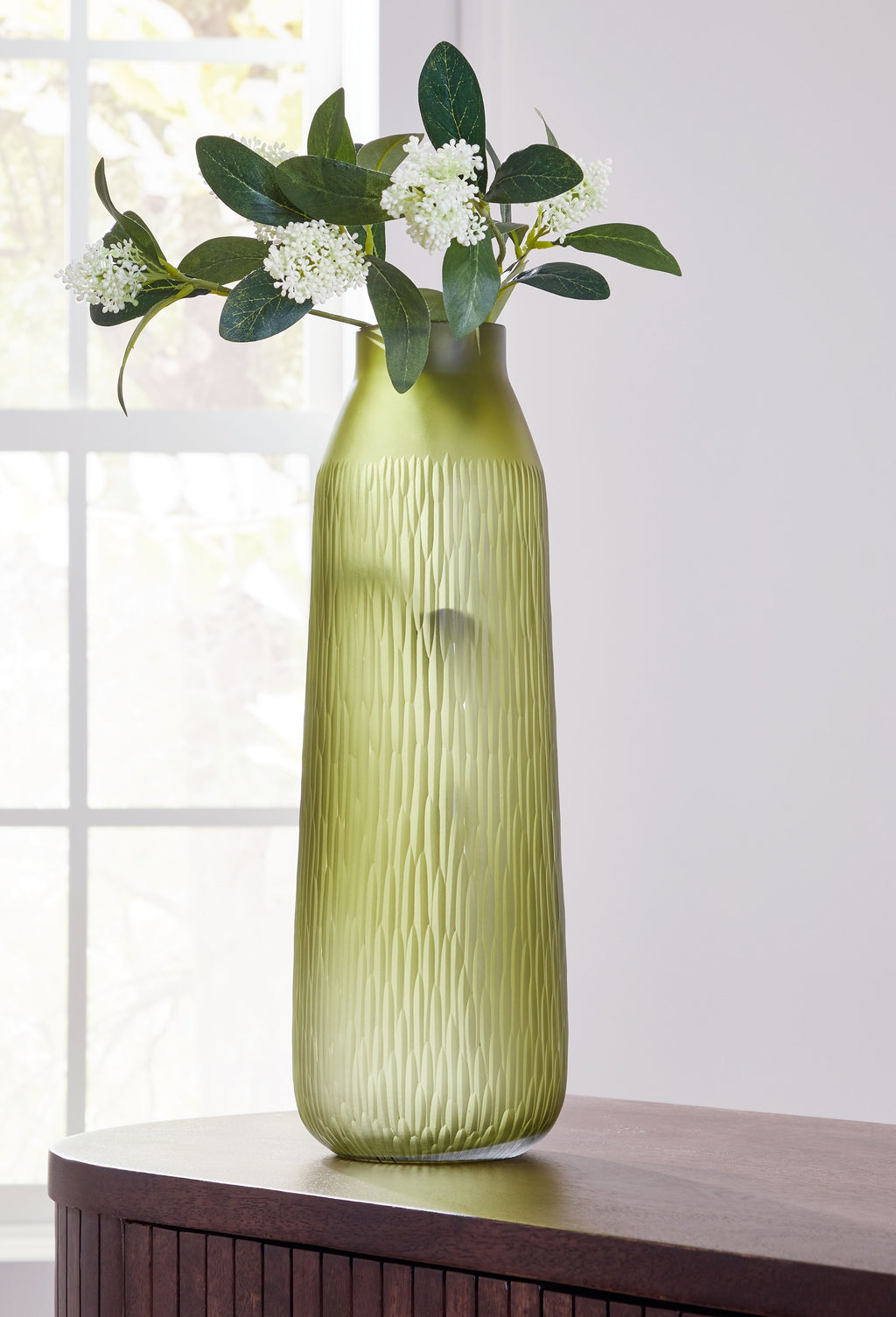 Green glass vase holding white blossoms and eucalyptus leaves with tall narrow silhouette.