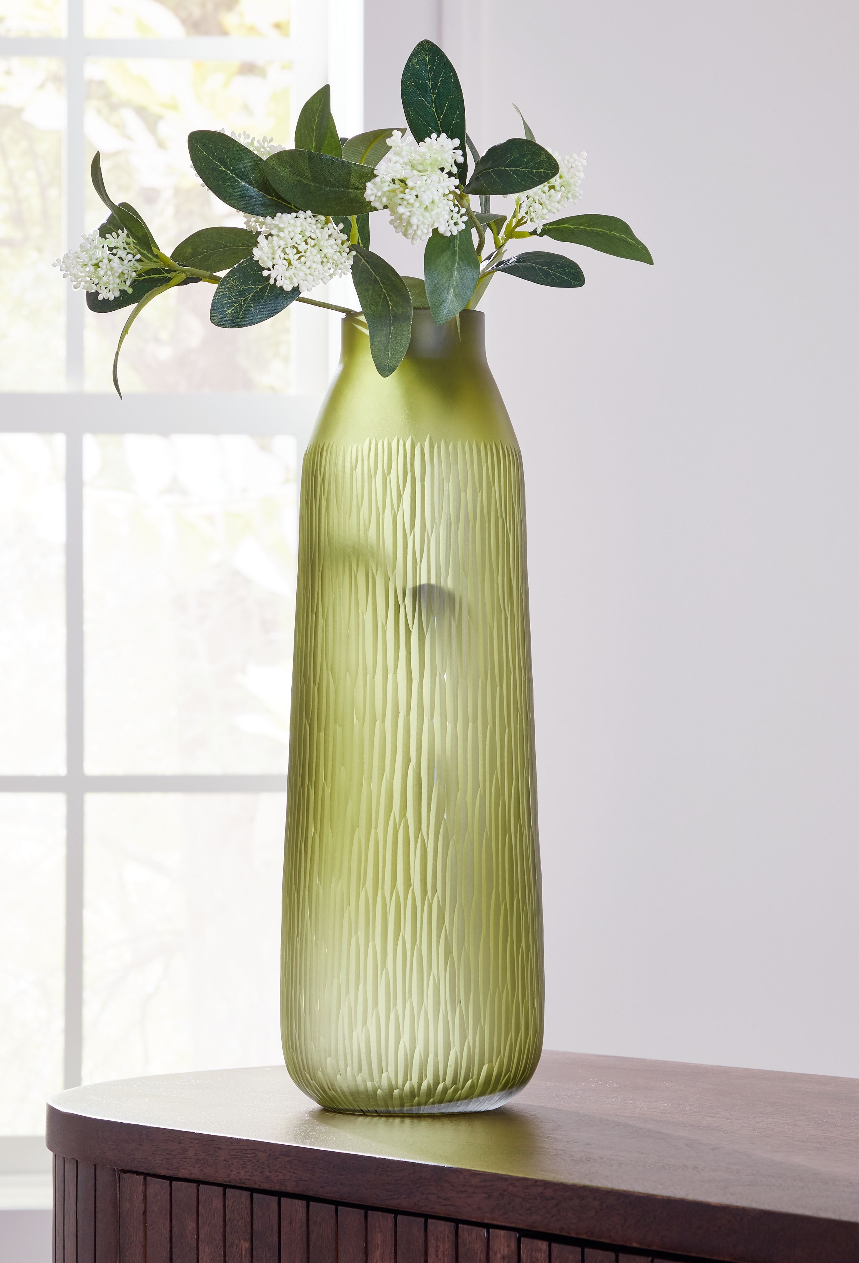 Green glass vase holding white blossoms and eucalyptus leaves with tall narrow silhouette.