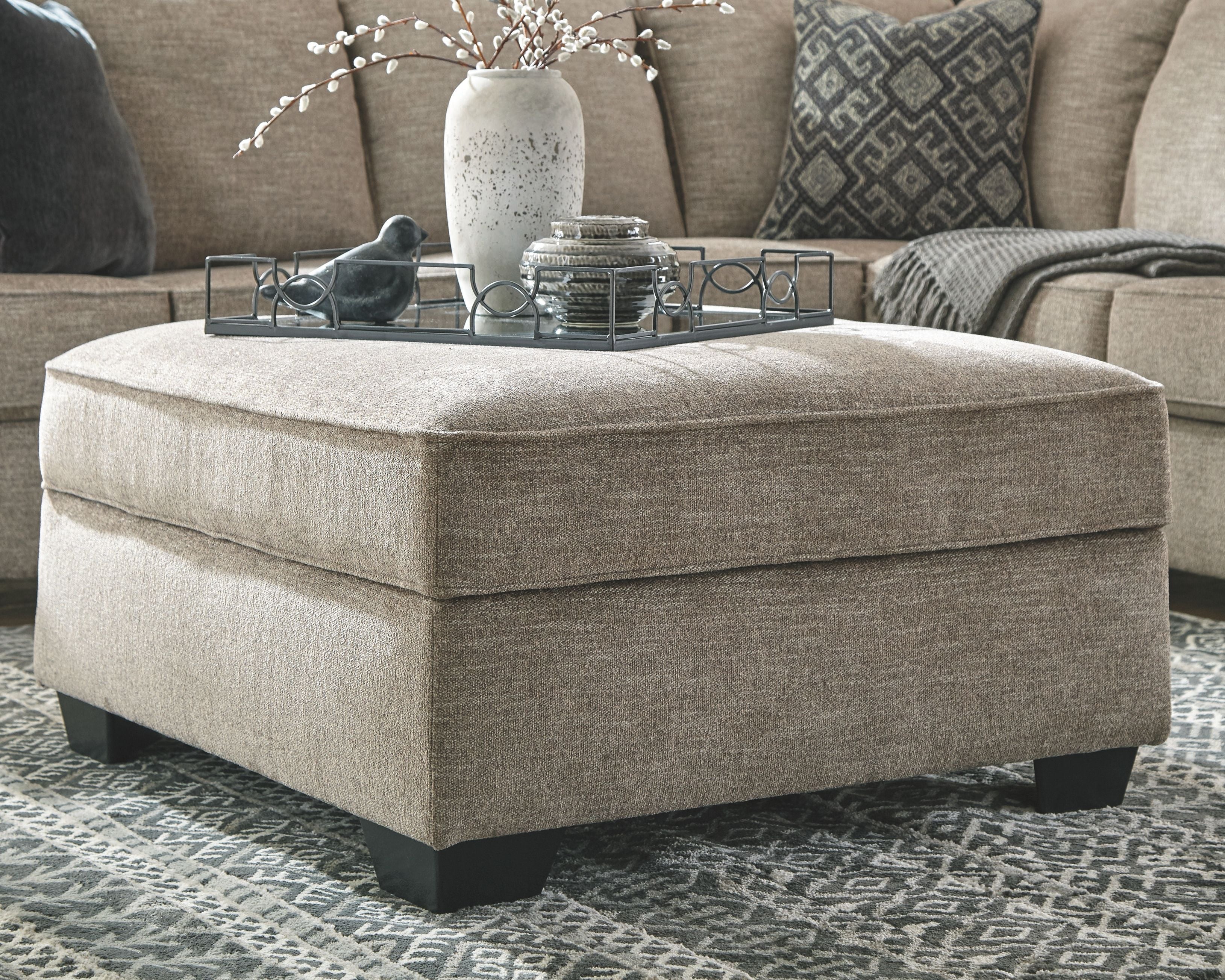 Beige ottoman with upholstered top and decorative tray on glass table