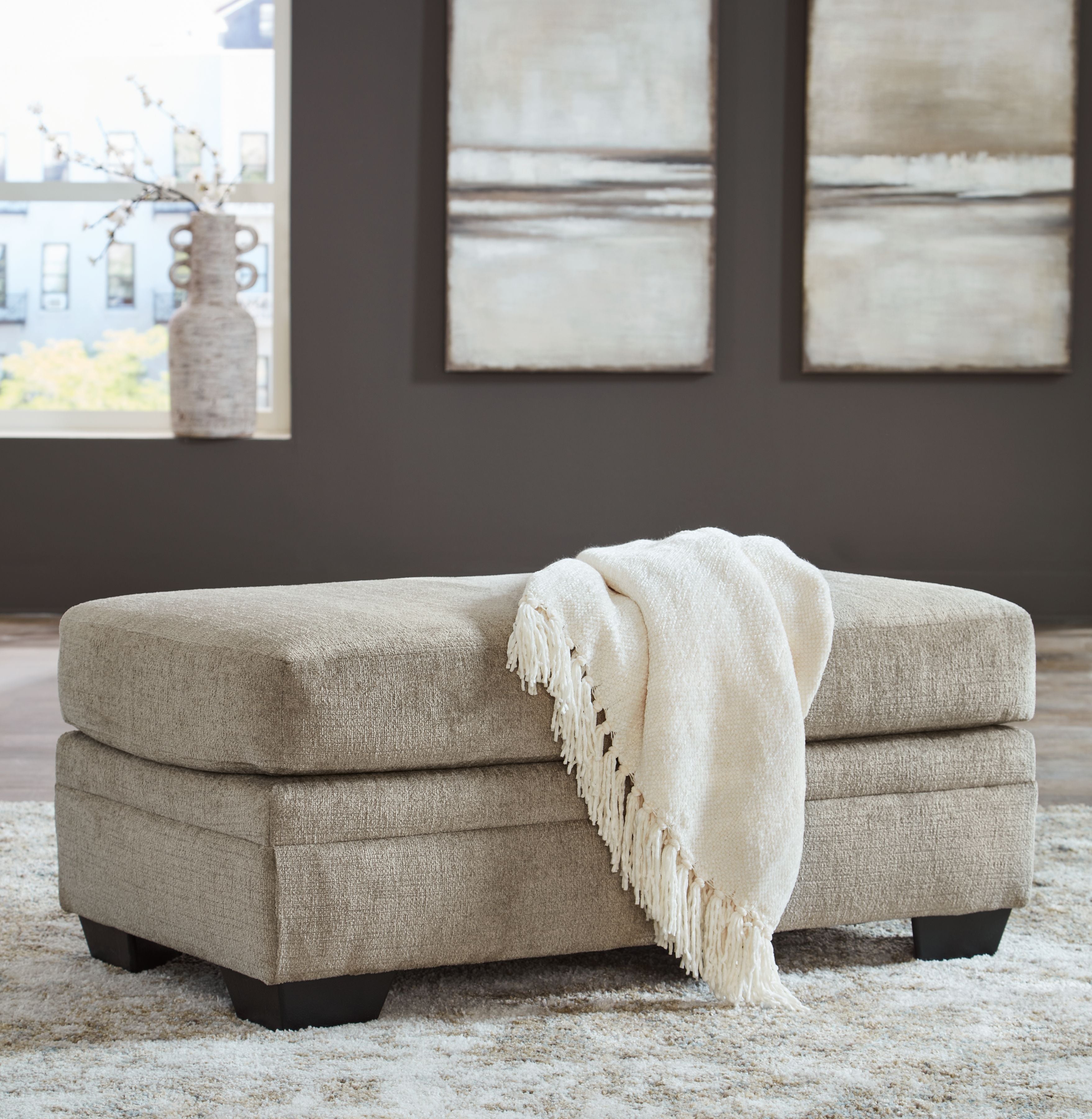 Upholstered ottoman in light beige with black legs and a folded throw on top.